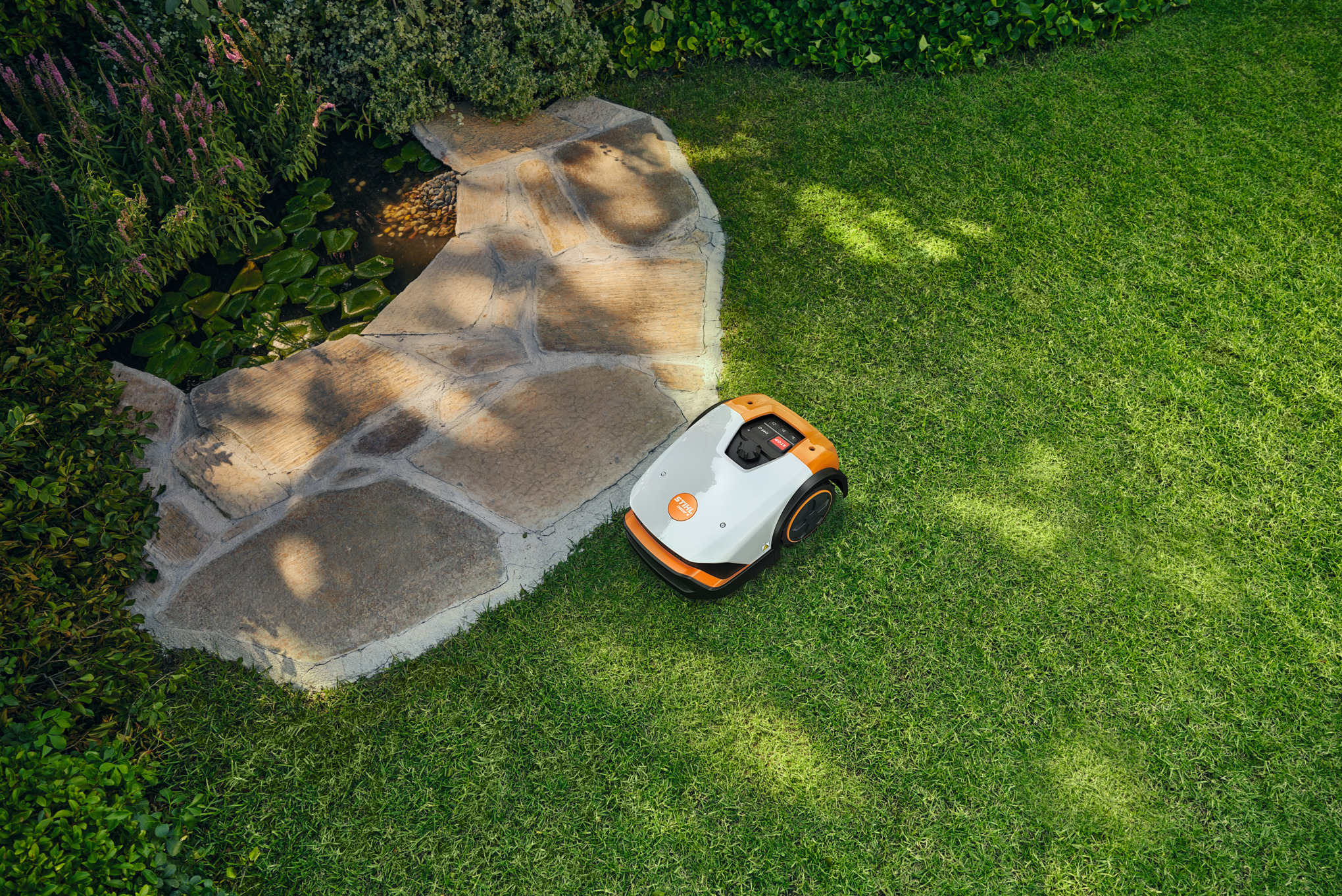 STIHL iMOW® 3 Mähroboter auf grünem Rasen vor einem gepflasterten Weg