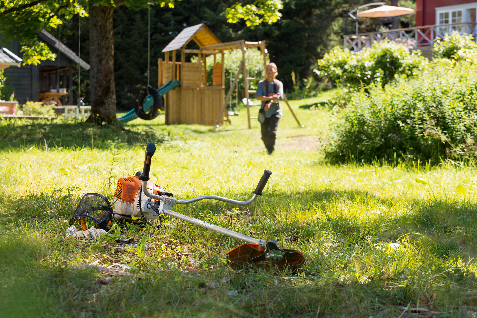 Motorsense STIHL FS 240 liegt im Gras, im Hintergrund bereitet eine Frau sich auf die Wartung vor 