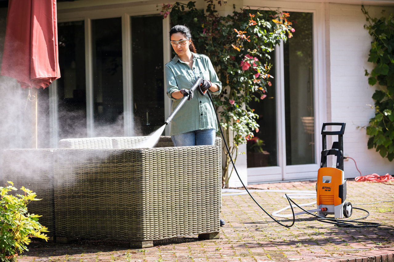 Frau mit Schutzbrille ist dabei, mit dem STIHL Hochdruckreiniger RE 88 auf der Terrasse ihre Gartenmöbel zu reinigen
